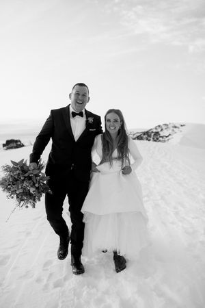 Black and white photo of a smiling bride and groom walking together in a snowy landscape on their wedding day.