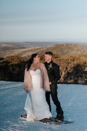 Bride in white gown with pink fur jacket and groom in black suit posing on snowy mountain slope at winter wedding.