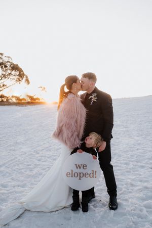 Couple kissing at sunset on snowy beach in wedding attire, child holding 'we eloped!' sign.