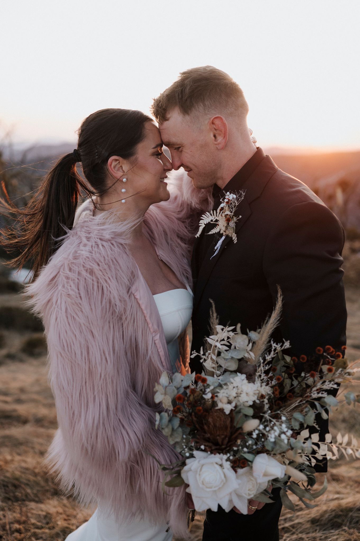 Couple sharing a romantic sunset moment, bride in white gown and pink fur wrap holding a boho bouquet, groom in black suit.