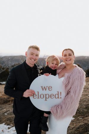 Couple holding a child and a round sign reading we eloped outdoors in winter attire, celebrating their elopement.