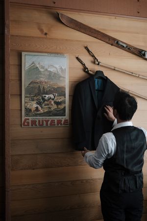 Man in vest arranging dark suit jacket on wooden wall near vintage Gruyere poster and antique skis.