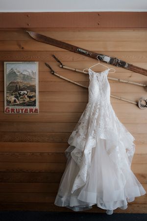 Elegant lace wedding dress hanging on rustic wooden wall with vintage Gruyere poster and antique skis.