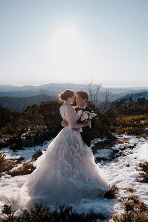 Bride and groom embrace on snowy mountain top, bride in lace gown, sun halo backdrop.