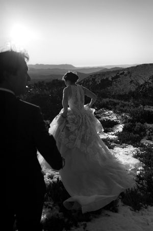 Black and white photo of a bride in a lace gown standing on a mountain overlook with groom at sunset.