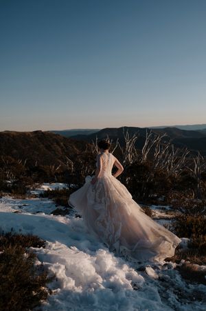 Bride in flowing blush gown stands on snowy mountain summit at sunset with vast landscape behind her.