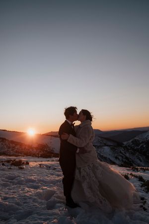 A couple shares a romantic kiss in wedding attire against a stunning mountain sunset on a snowy landscape.