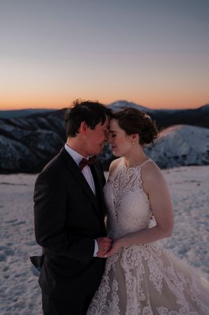 Couple in wedding attire sharing romantic moment on snowy mountain peak at sunset with orange sky backdrop.
