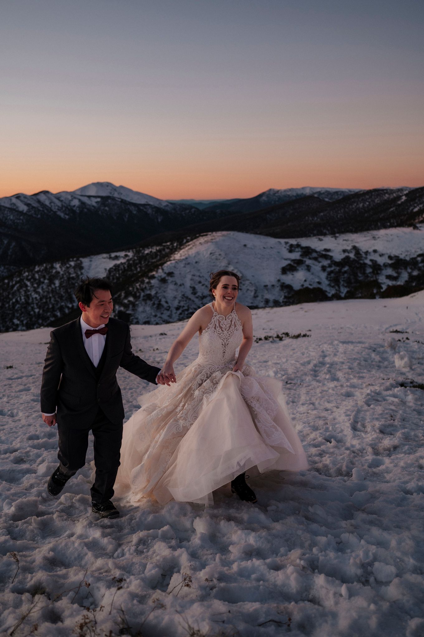 Bride and groom walk hand-in-hand through snowy mountain landscape at sunset during winter wedding photo session.
