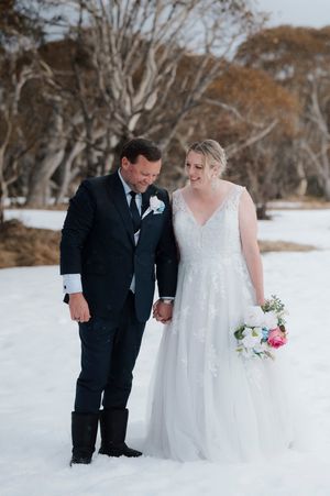 Bride and groom holding hands smiling in snowy winter wonderland wedding setting with bare trees.