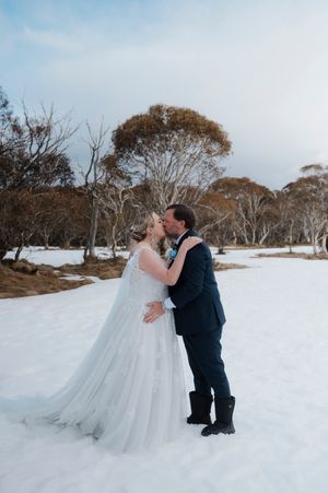 Bride and groom sharing romantic kiss in snowy outdoor winter wedding with bare trees backdrop.