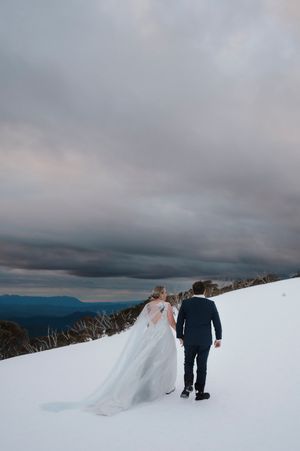Bride and groom walking hand in hand through snowy mountain landscape under dramatic cloudy sky.