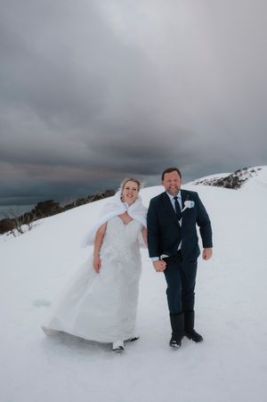 Bride and groom holding hands walking in snow on their winter wedding day under dramatic cloudy skies.