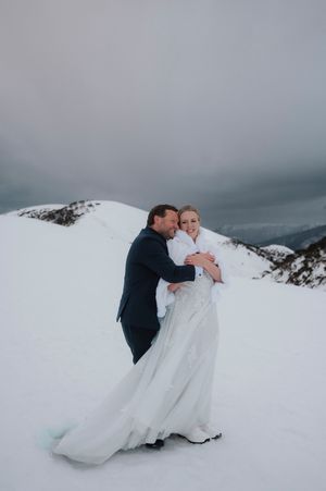 Bride and groom embracing on a snowy mountain peak, bride in white gown with fur wrap, groom in navy suit.