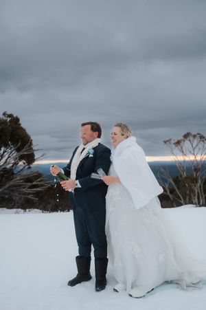 Bride and groom celebrate in snowy mountain landscape, she wears white fur wrap over wedding gown under dramatic winter sky.
