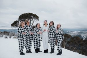 Bride in white dress with bridesmaids in matching black and white plaid jumpsuits celebrating on a snowy mountain.