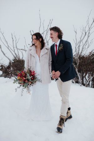 Bride and groom walking hand in hand through snow, she holds a red bouquet wearing a fur coat over her wedding dress.