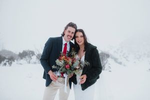 Smiling newlywed couple posing together in snowy winter landscape, groom in navy suit with red tie, bride holding bouquet.