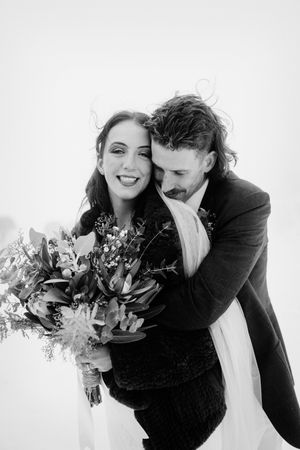 Black and white wedding photo of smiling bride holding bouquet while groom embraces her from behind outdoors.