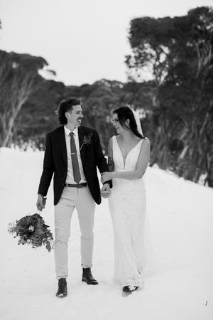 Black and white photo of a bride and groom walking hand in hand through snow, smiling at each other on their wedding day.