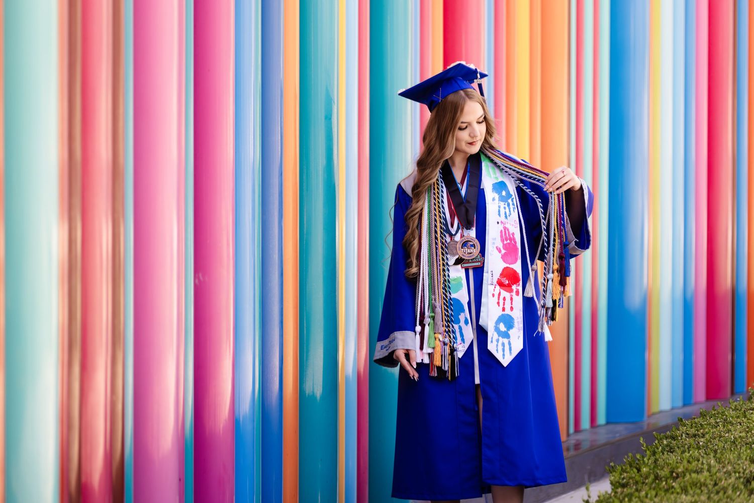 Young woman in blue graduation cap and gown poses proudly before a vibrant colorful striped wall holding books.
