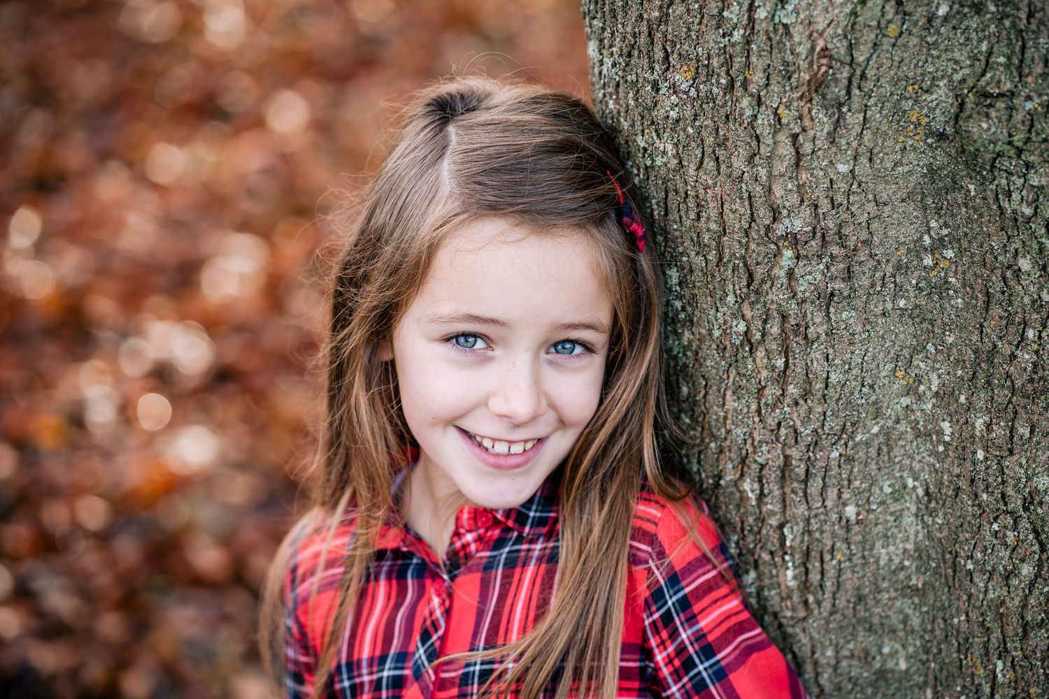 Smiling young girl with blue eyes and long brown hair leaning against a tree in a red plaid shirt during autumn.