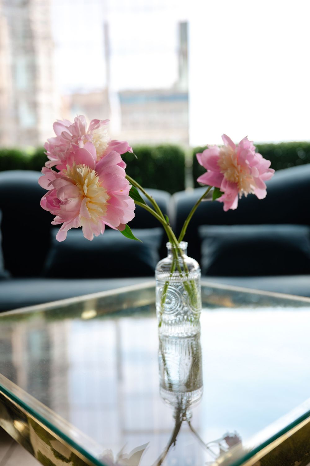 Pink peonies in a clear glass vase on a gold mirrored coffee table with a dark sofa in the background.