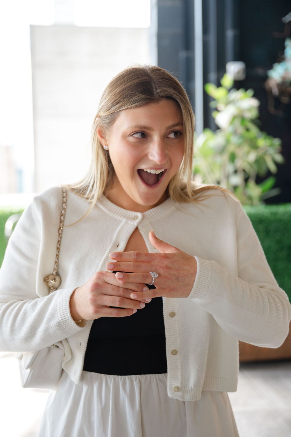 Excited blonde woman in white outfit displaying engagement ring with surprised expression.
