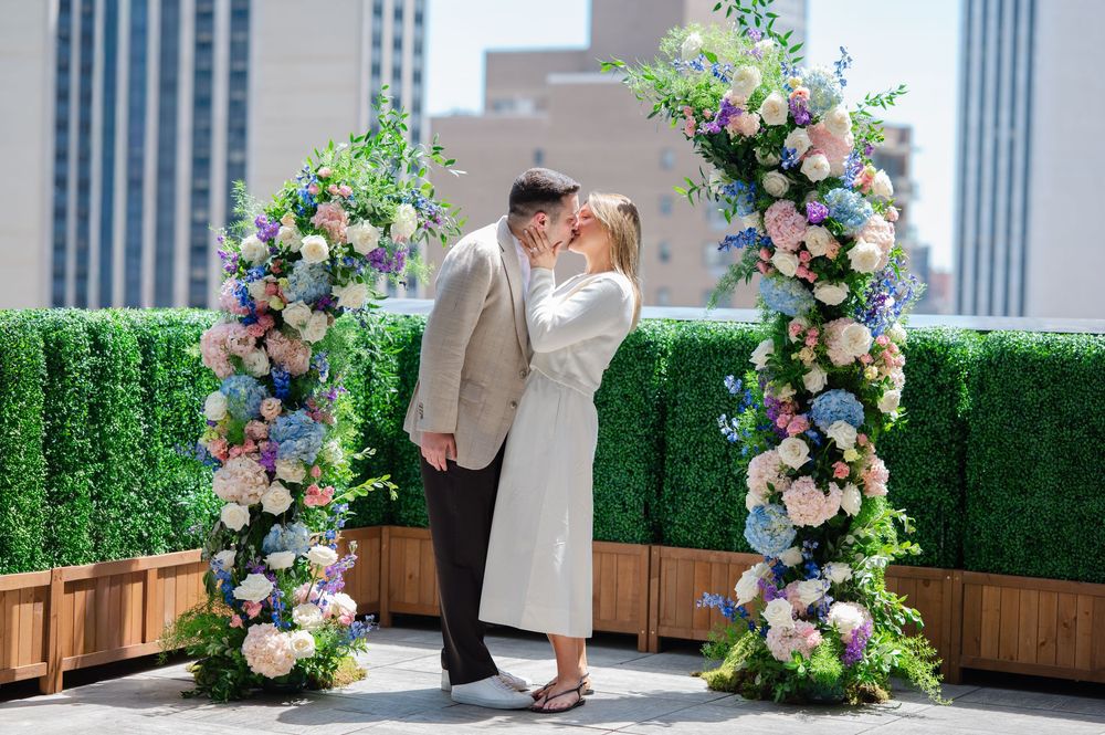 Couple kissing under floral arch with pink, blue, and white flowers on a rooftop wedding venue.