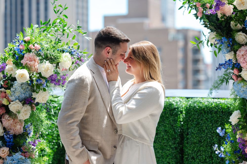 Couple sharing romantic moment near colorful floral arch during rooftop proposal ceremony.