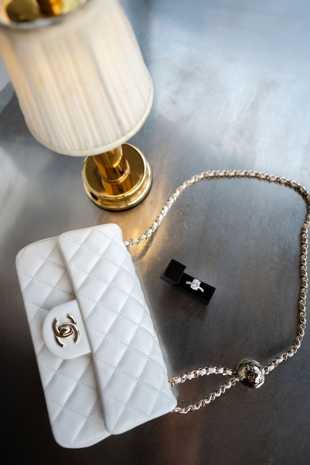 White quilted Chanel mini flap bag with gold chain strap beside a ring box and small lamp on table.