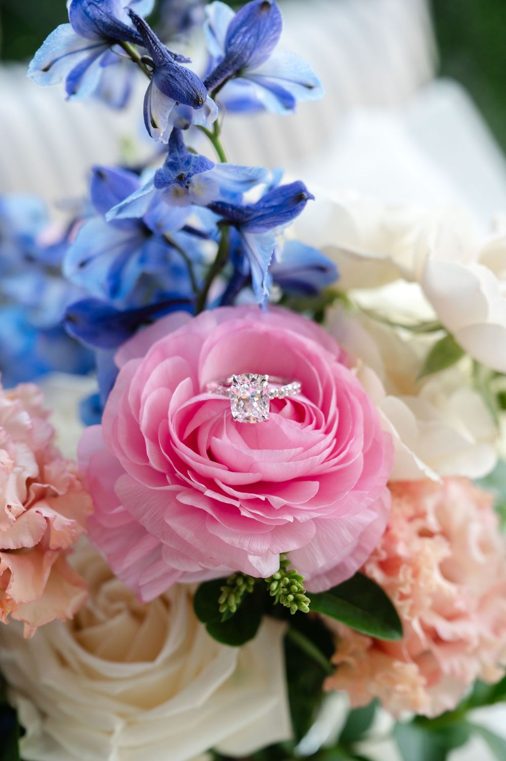 Diamond engagement ring resting on a pink rose surrounded by blue, white, and peach wedding flowers.
