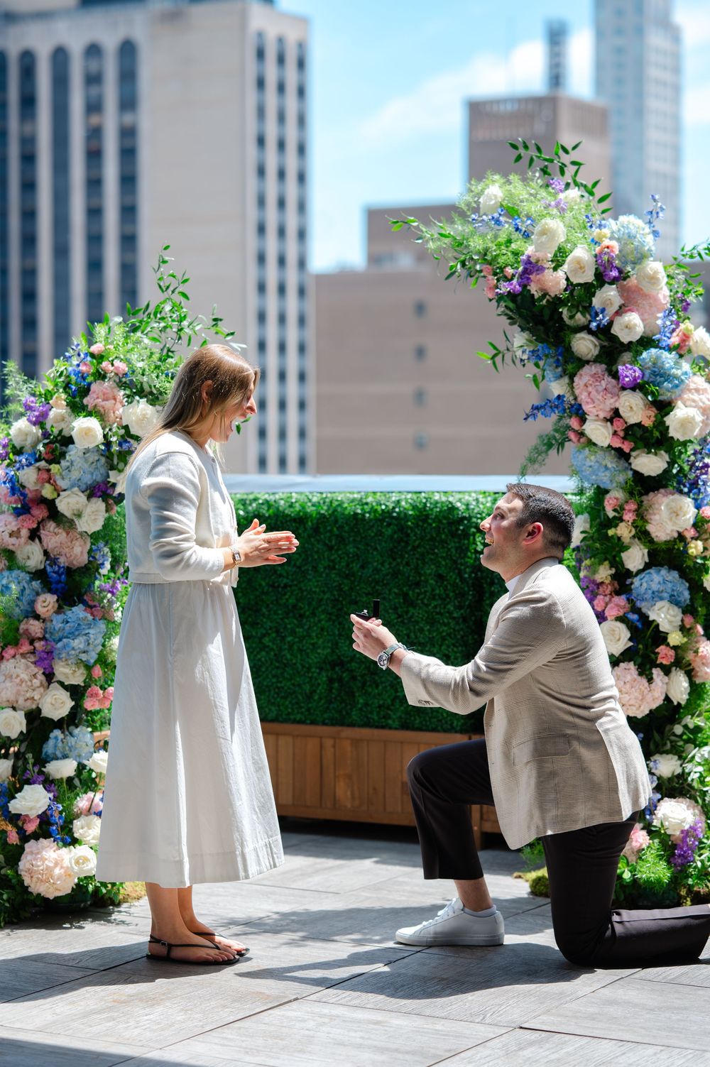 Man proposing on one knee to woman in white dress at a floral arch on a NYC rooftop.