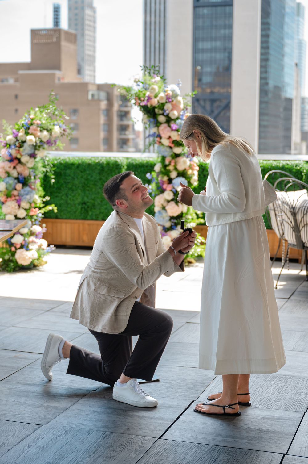 Man proposes on one knee to woman on rooftop terrace with floral arch and city skyline backdrop.