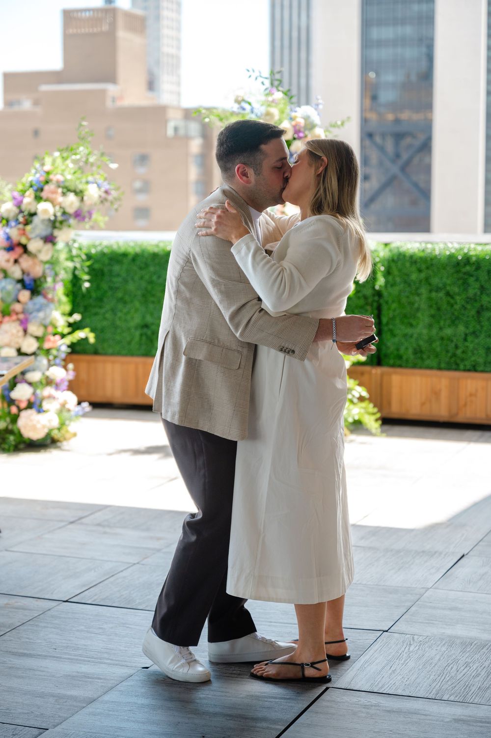Couple sharing a romantic kiss on a rooftop venue with floral arrangements and city skyline backdrop.