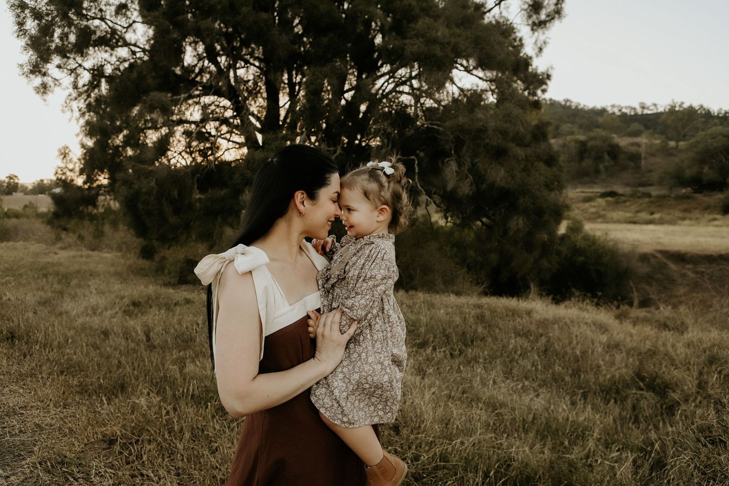A mother holds her smiling toddler in a sunlit field, sharing a tender nose-to-nose moment with trees in the background.