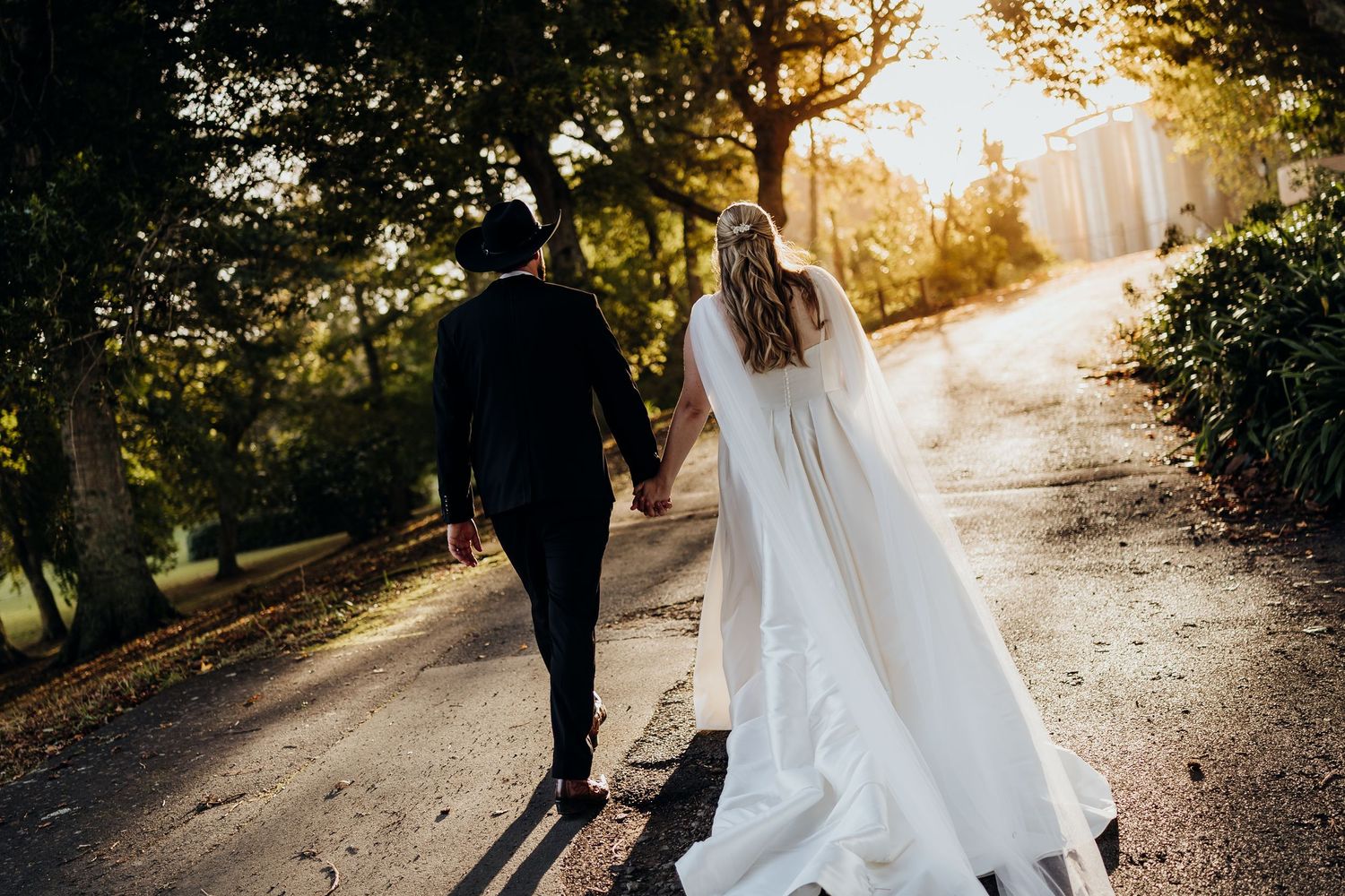 A couple in traditional Indian wedding attire walks through a forest path at sunset.