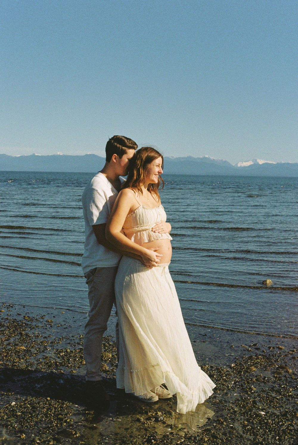 Couple embracing on rocky beach, pregnant woman in white dress, mountains and blue water backdrop.