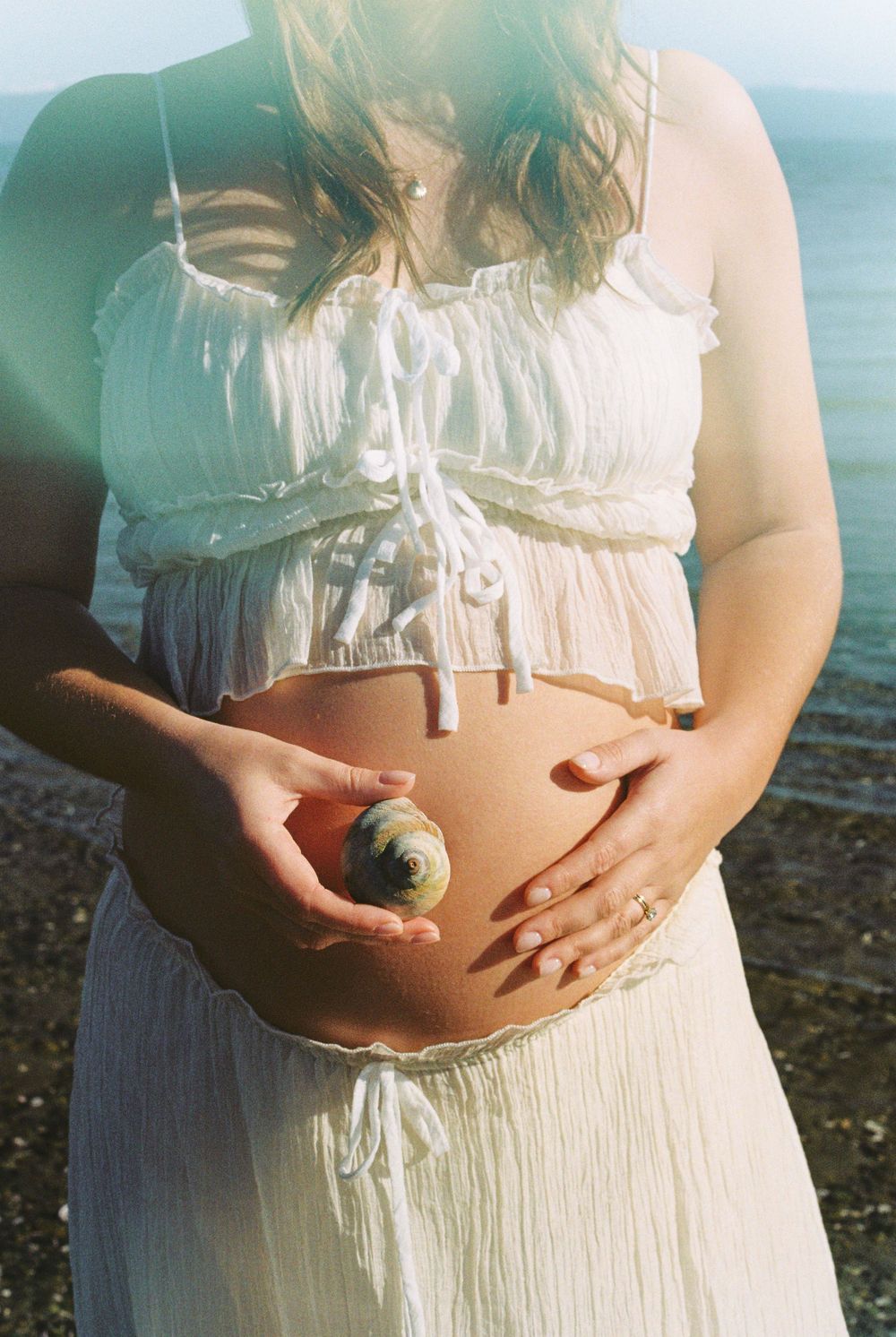 Pregnant woman in white dress holding a seashell against her bare baby bump at a sunny beach.