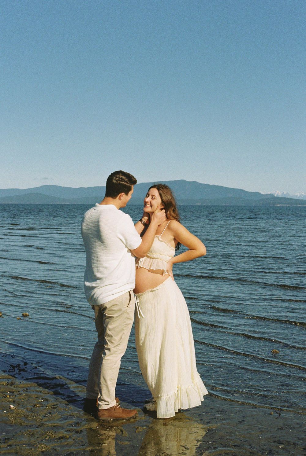 Couple sharing tender moment standing in lake shallows, pregnant woman in white dress smiling at partner.