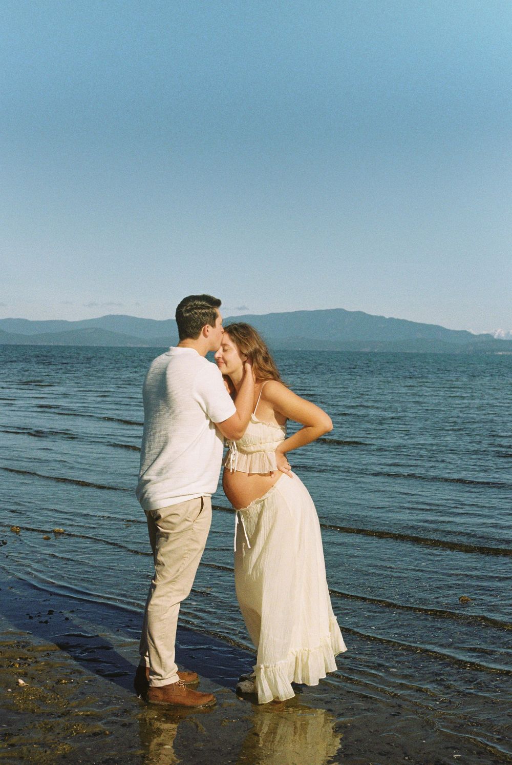 Couple sharing an intimate kiss on a scenic beach during a golden hour maternity shoot.