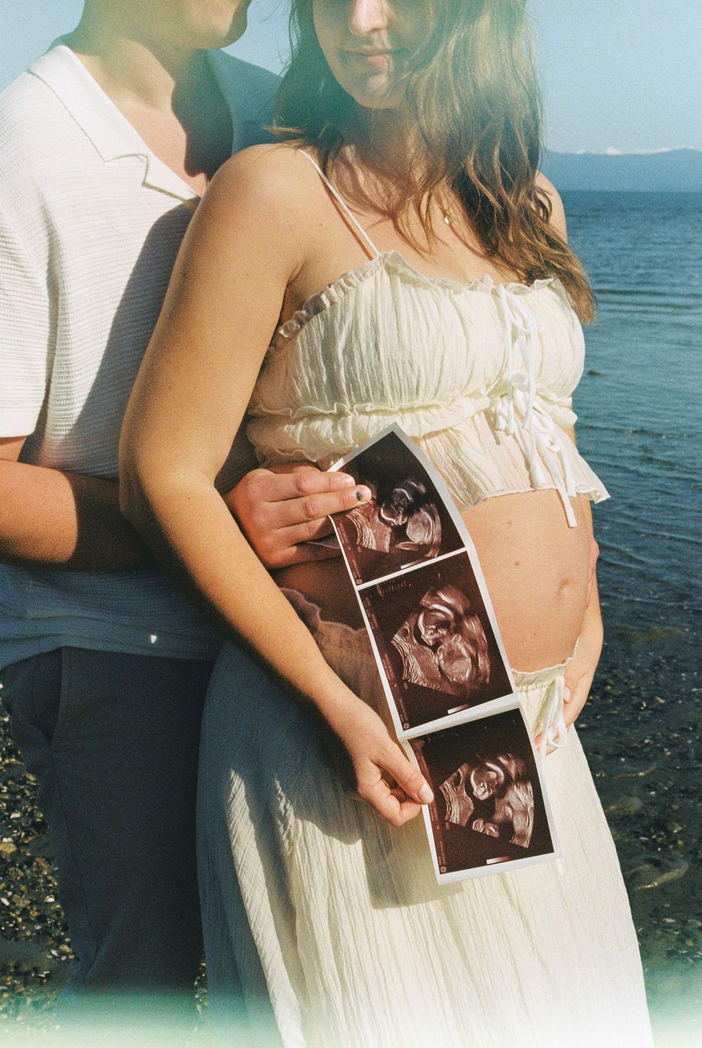 Pregnant couple holding ultrasound photos near a lake, woman in white dress showing baby bump outdoors.