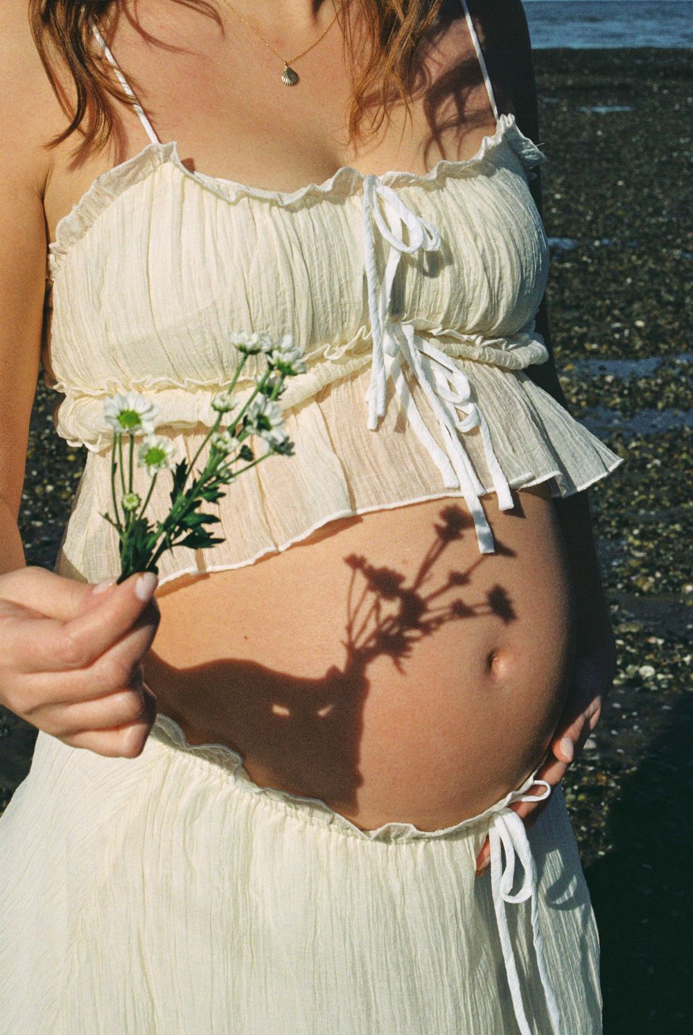 Pregnant woman in white crop top holding wildflowers, baby bump visible with flower shadows, standing near rocky beach.