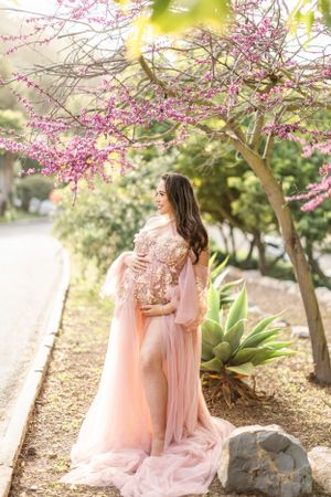 A romantic maternity photoshoot under blooming pink cherry blossoms with a flowing peach gown in a garden setting.