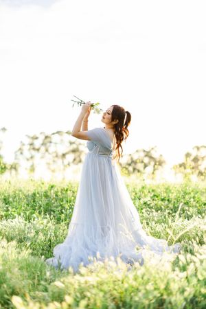 A person in a flowing white dress poses with flowers in a sunlit meadow.