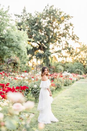 Woman in white dress poses elegantly among red roses in garden setting.