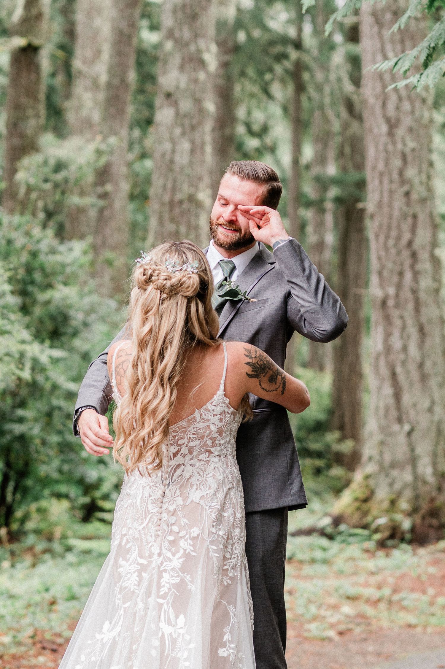 Emotional groom wiping tears as bride in lace wedding dress turns around for first look in forest. Photo by Bremerton Wedding Phototographer
