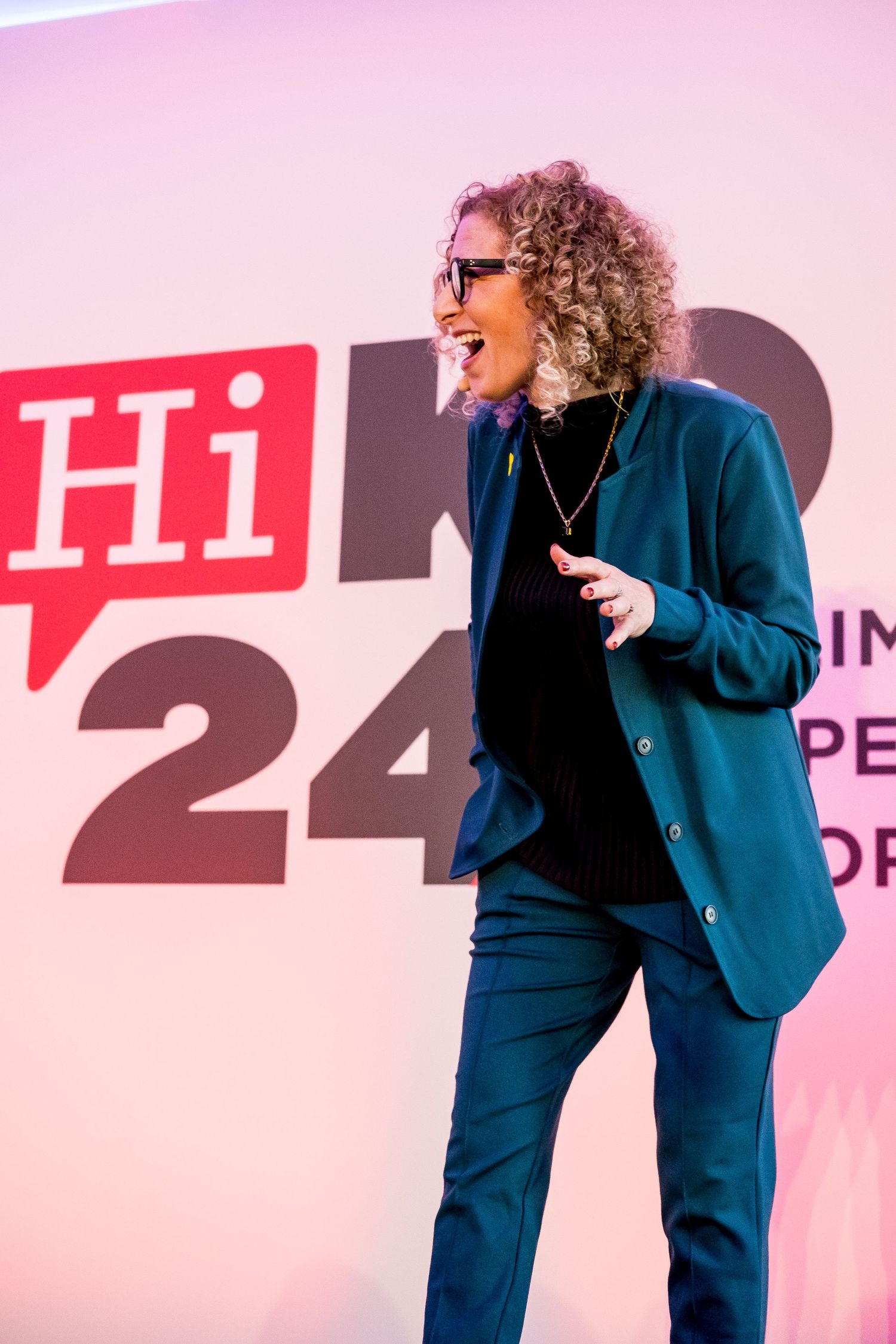 A curly-haired woman in teal blazer laughing on stage at HiRo 24 conference event.