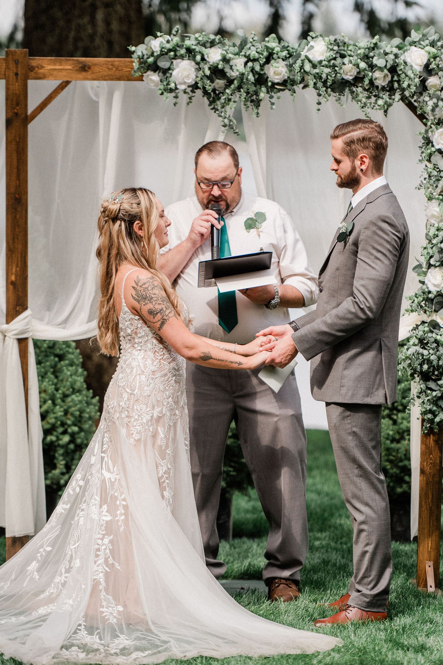 Outdoor wedding ceremony with bride and groom exchanging vows under a floral arch with white draping and greenery. Photo by Pine + Vow