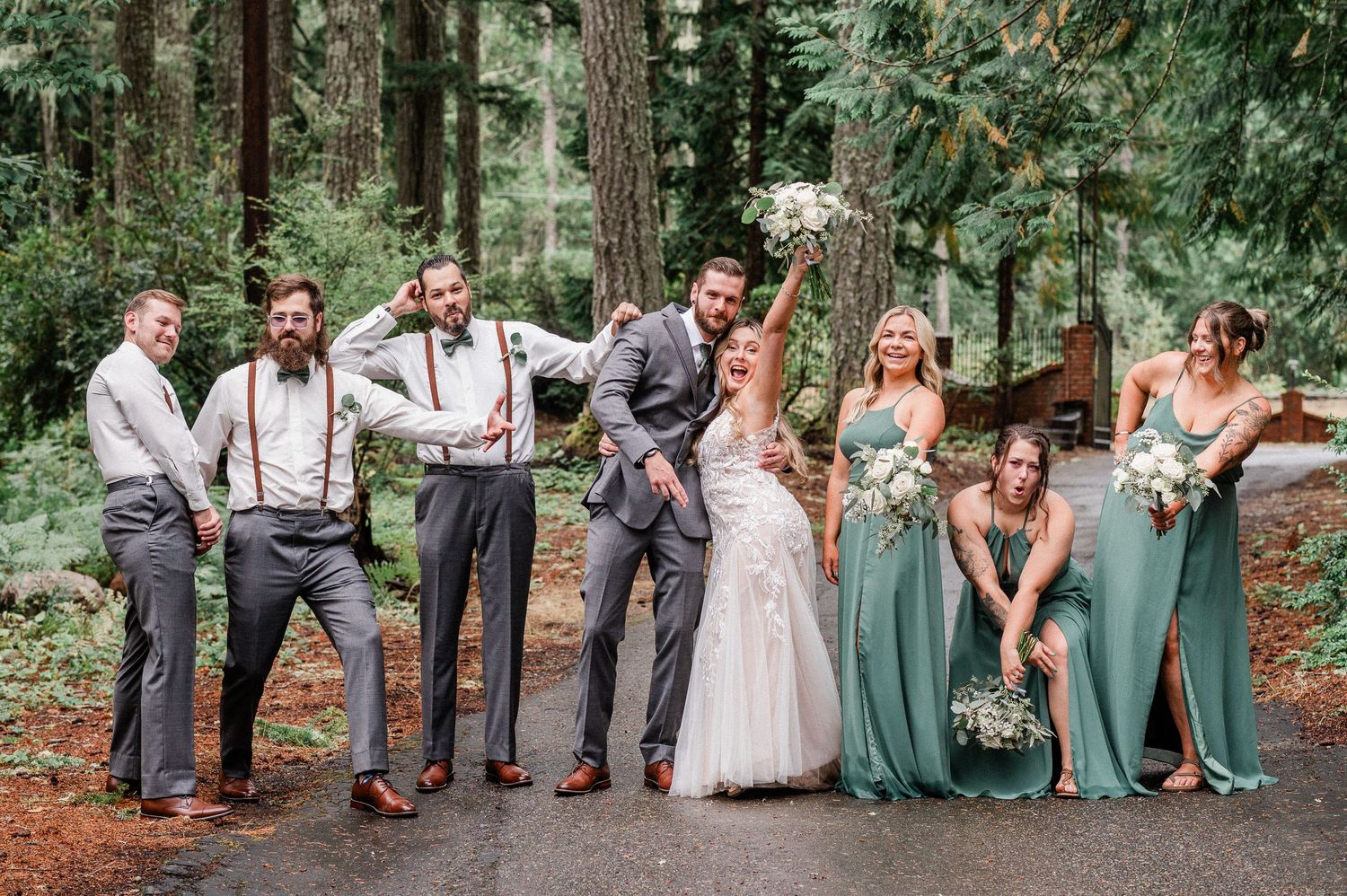 Wedding party posing playfully in a forest setting, groomsmen in gray suits and bridesmaids in sage green gowns.
